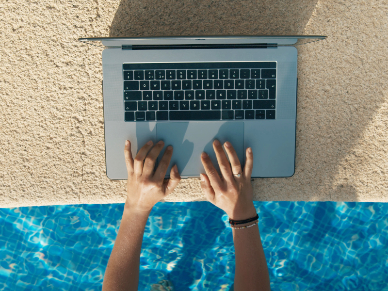 Fotografia przedstawia osobę, która korzysta z laptopa, będąc w basenie. Komputer ustawiony jest na brzegu basenu. Widoczne są ręce osoby go obsługującej.